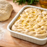 L’impasto della focaccia prende forma: morbido, vivo, pronto a lievitare lentamente. Nella teglia si intravedono già le classiche fossette della focaccia, dove l’olio extravergine d’oliva si raccoglie e profuma l’impasto. Accanto, il pane in attesa e la farina sul tavolo raccontano una cucina fatta di gesti semplici e ingredienti autentici, proprio come nella tradizione contadina.